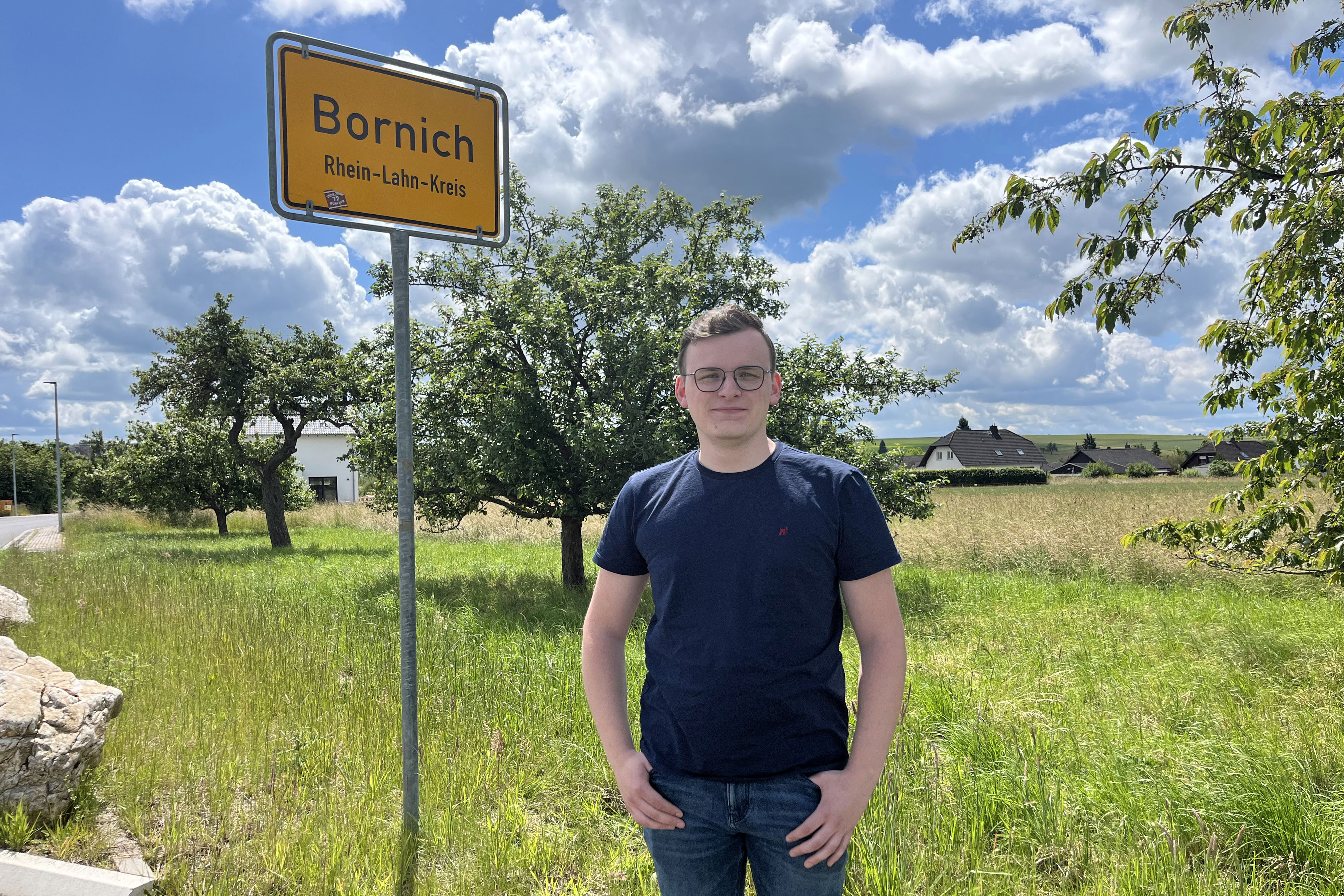 Ein junger Mann steht vor einem Ortseingangsschild mit Aufschrift Bornich
