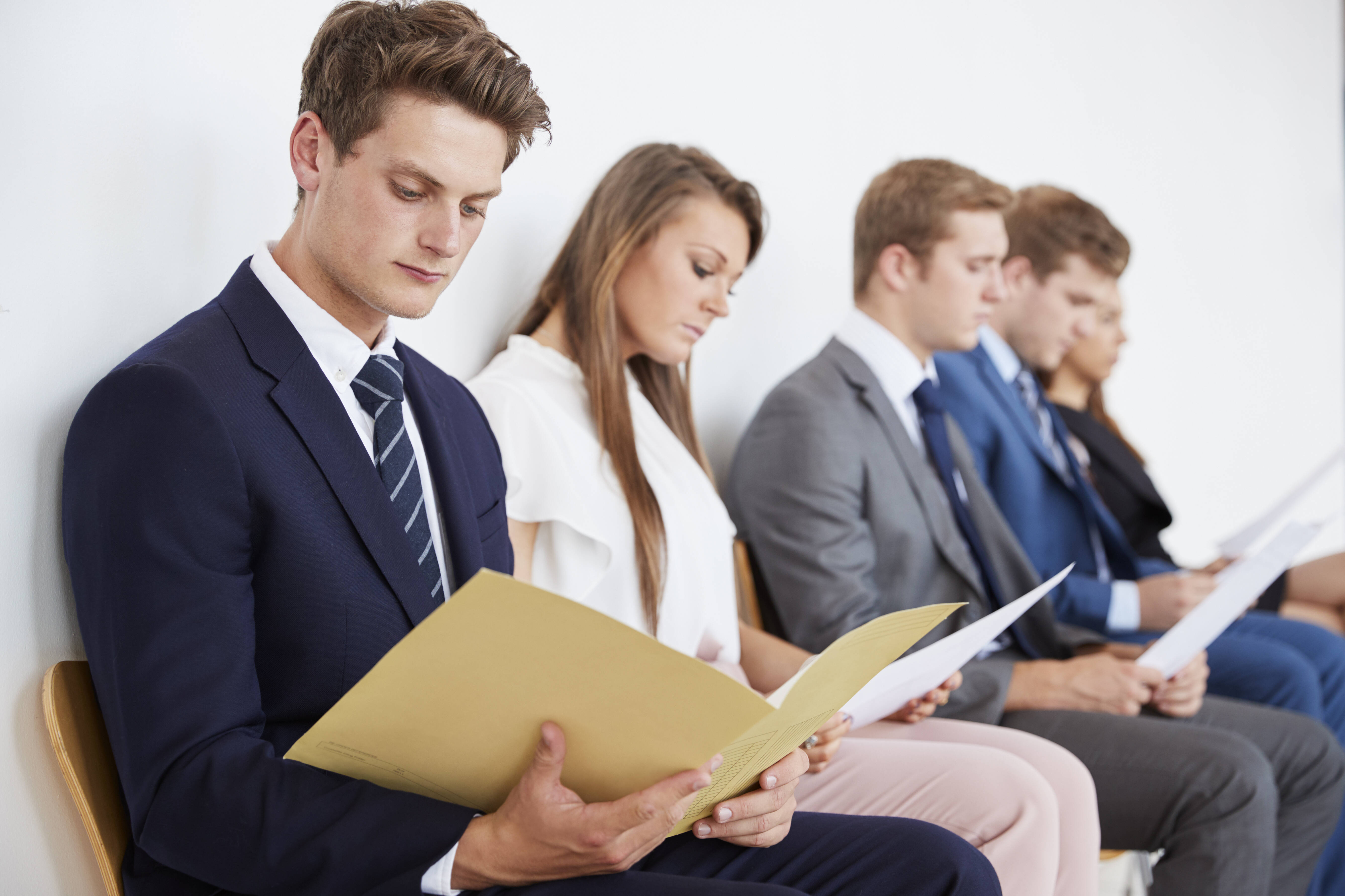 Junge Menschen in Anzügen sitzen in einer Reihe mit Bewerbungsmappen in der Hand